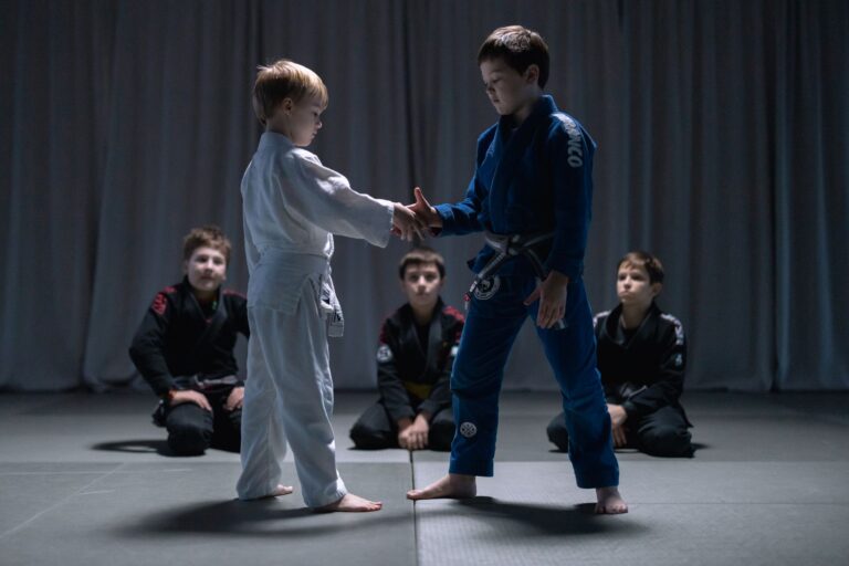 Two young boys in gis shaking hands during a jiu jitsu practice in a gym setting, under dim lighting.