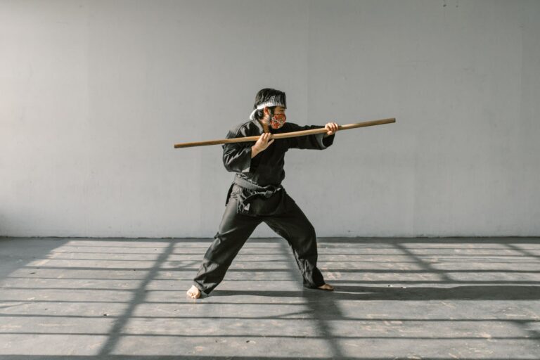 Martial artist in black uniform demonstrates skill with a wooden staff indoors.