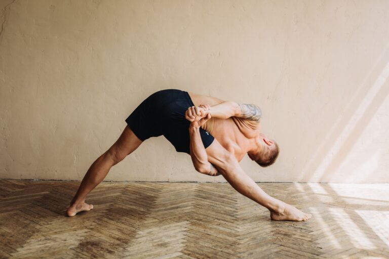 Athletic man performing a deep stretching yoga pose indoors with bare feet and tattoos.