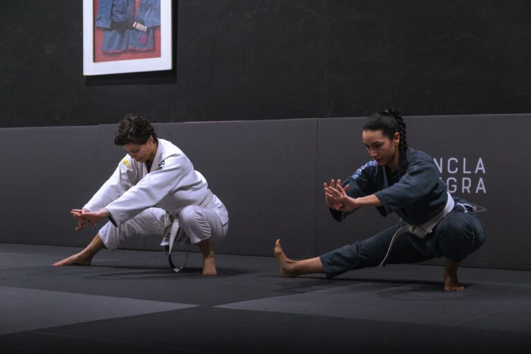 Two women in Brazilian Jiu-Jitsu attire practicing stretches on a mat indoors.