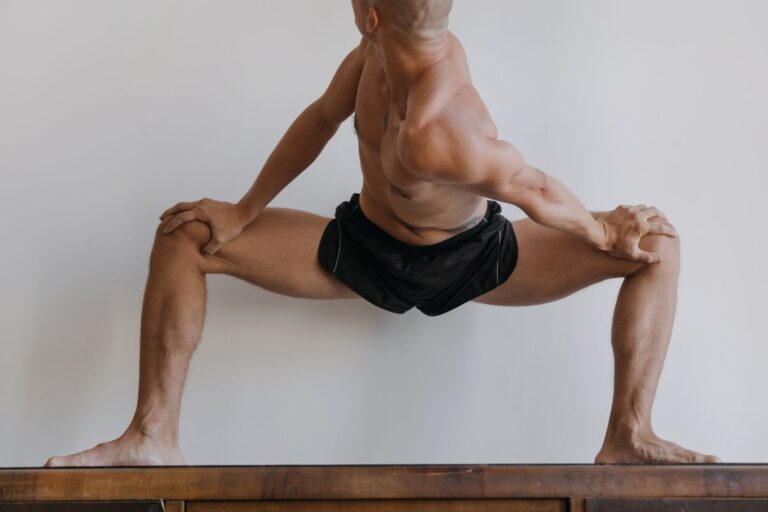A shirtless man with a muscular build stretches on a wooden surface indoors.