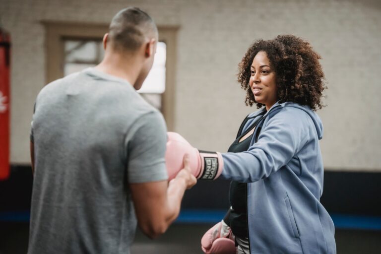 Positive overweight female in sportswear putting on gloves with support of personal coach before boxing workout